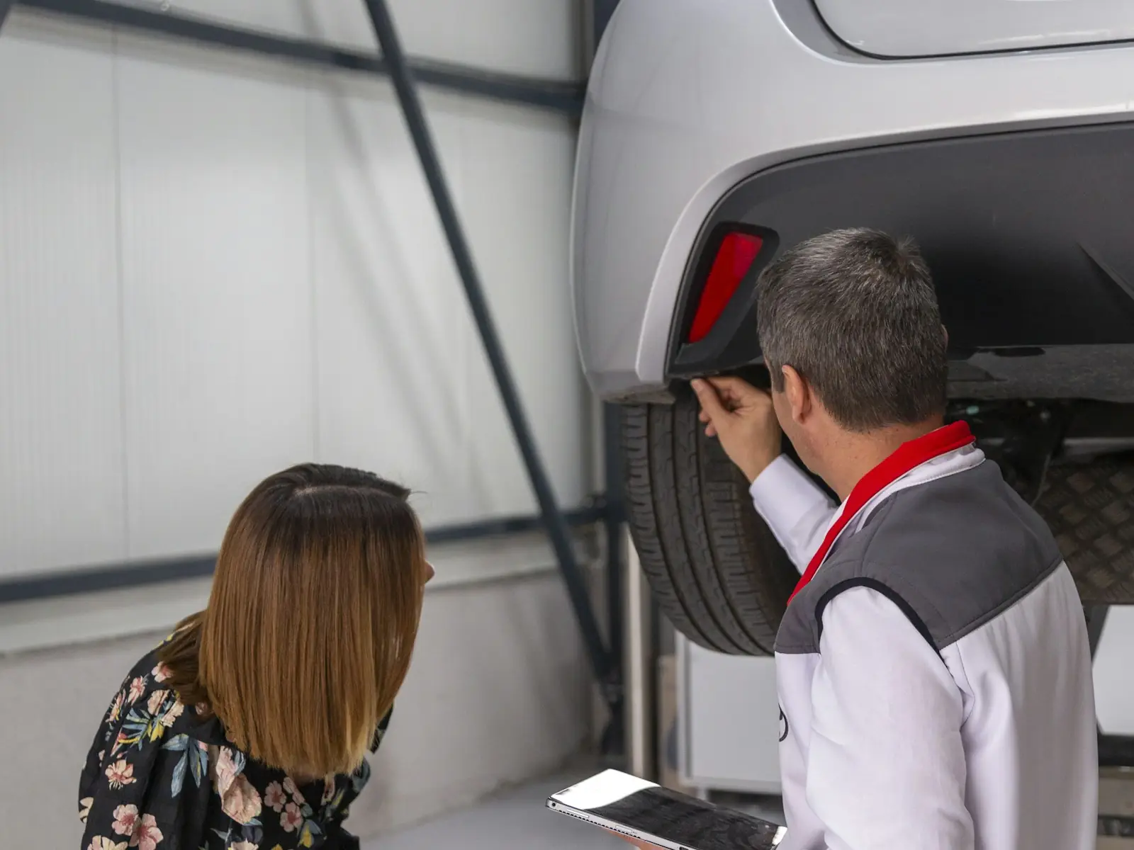 A mechanic with a tablet checks under a raised car while the owner looks on